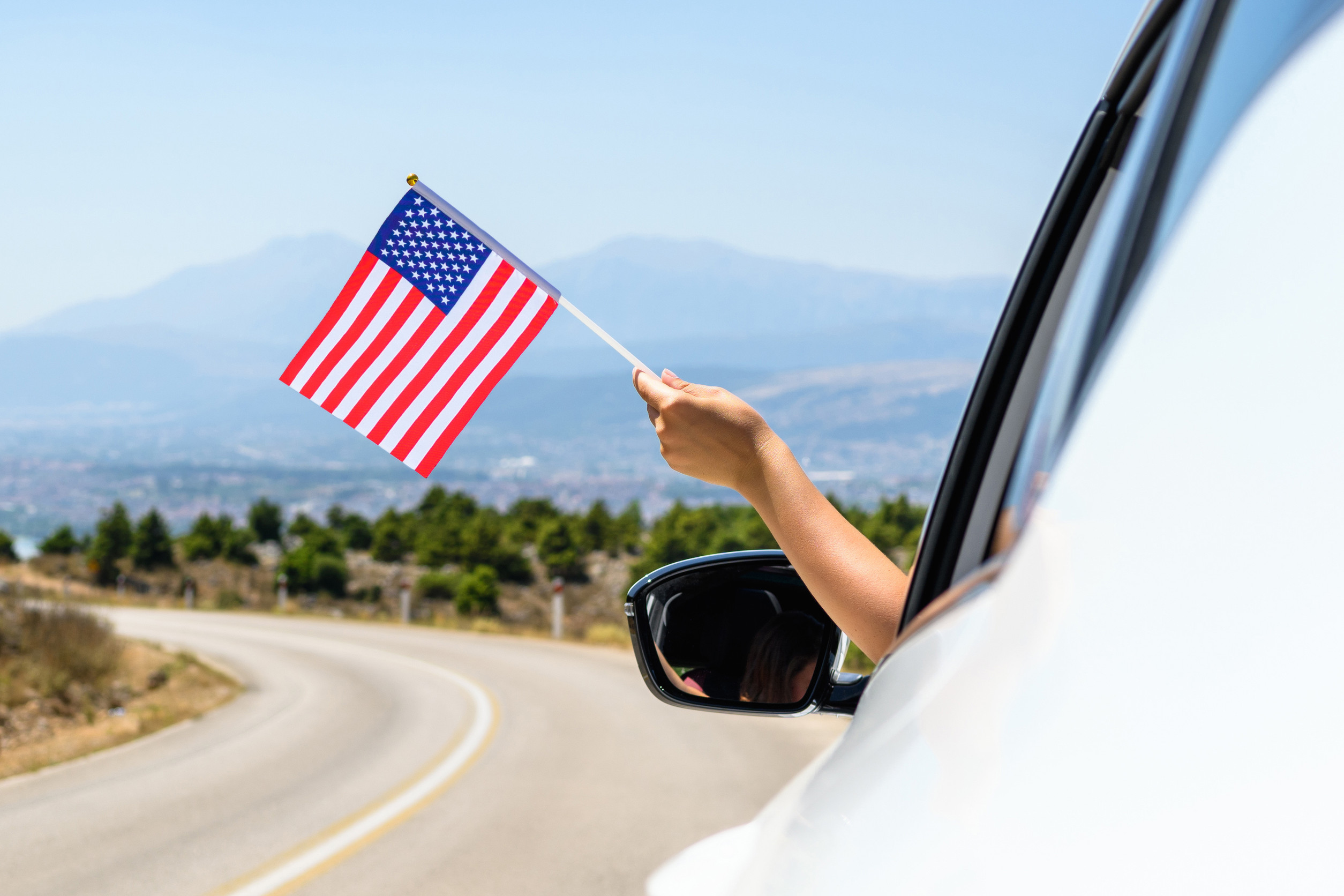 Holding USA flag from the open car window driving along the serpentine road in the mountains.