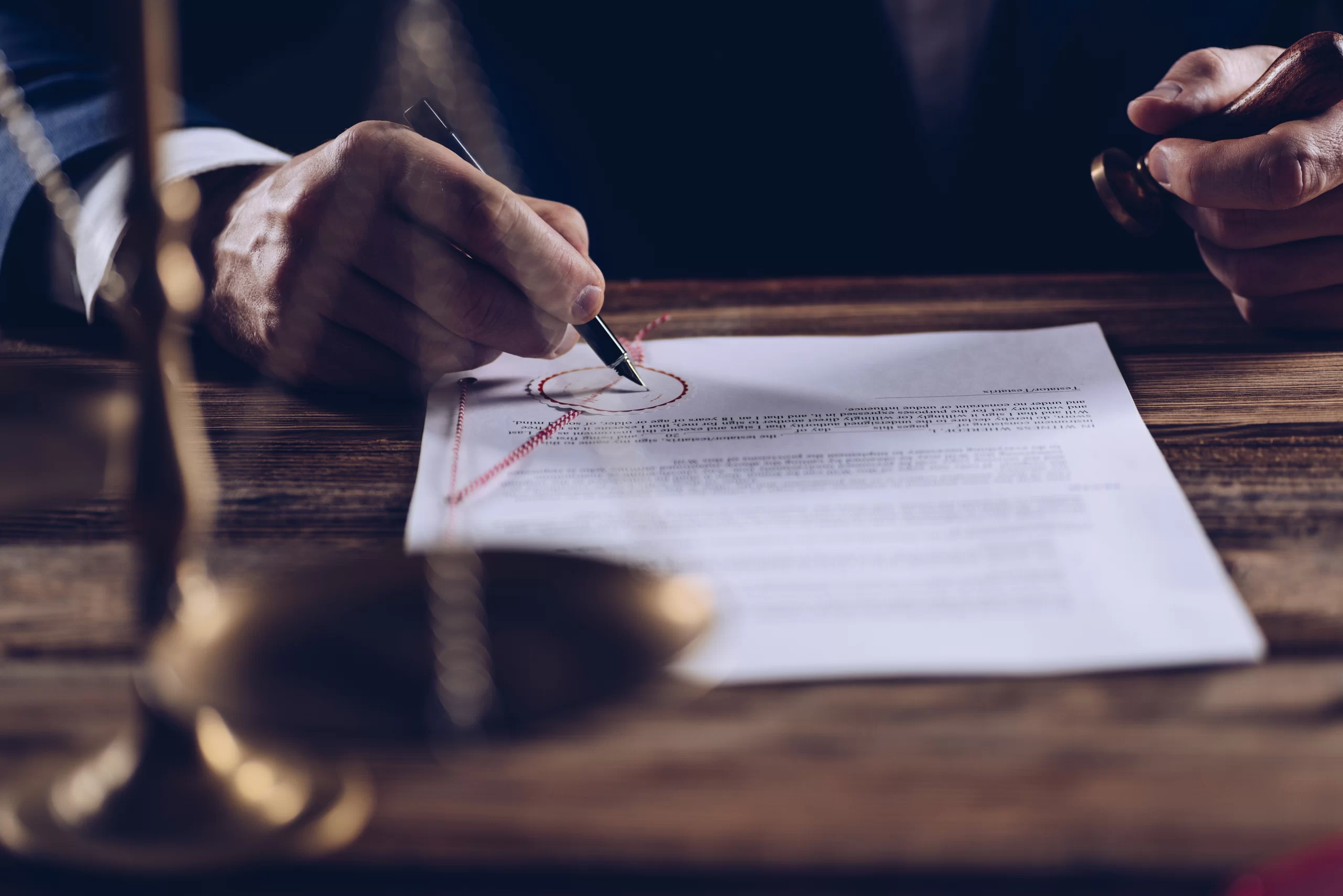 Close-up of a lawyer signing and stamping a legal document at a desk with justice scales in the foreground.