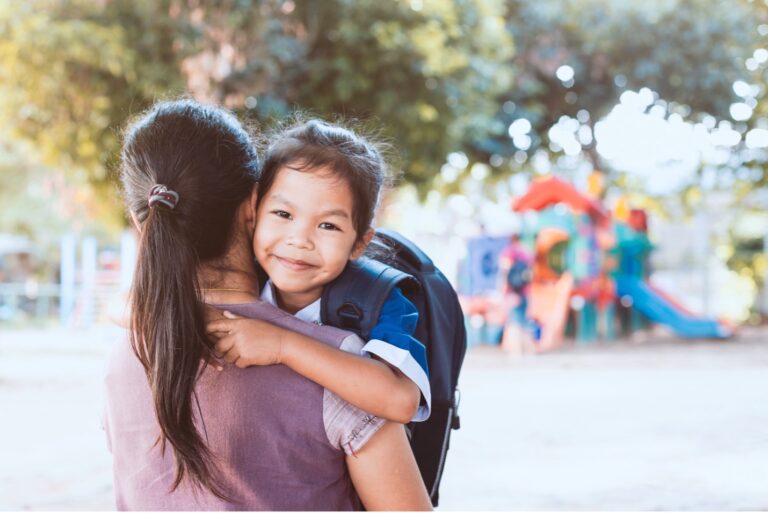 Young child smiling and hugging their parent outside a school or daycare, wearing a backpack.