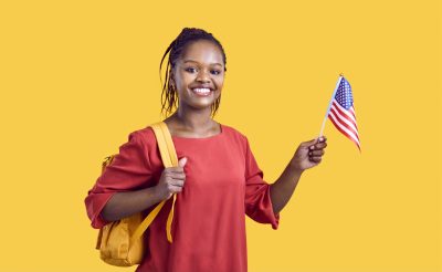 Smiling young woman wearing a backpack and holding a small American flag, standing against a bright yellow background.